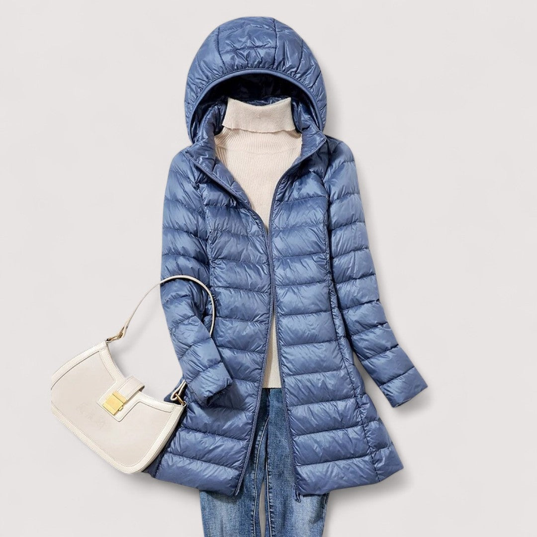 Blue hooded longline puffer jacket worn over a cream high-neck sweater and jeans, paired with a white handbag.