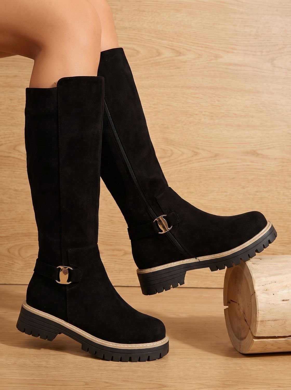 Black knee-high boots displayed at an angle, showing side zippers, decorative buckles, low block heels, and rugged lug soles.