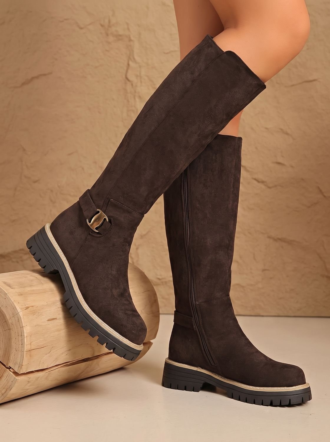 Brown knee-high boots posed with one elevated on a log, clearly showing the tall silhouette, side buckle, and heavy lug tread.