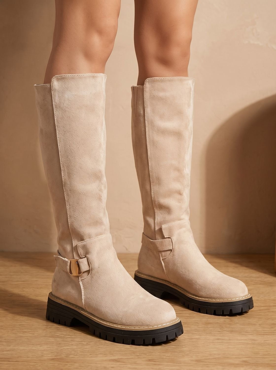 Cream knee-high boots modeled standing on a wooden floor, showing their tall shafts, rounded toes, side buckles, and thick tread.