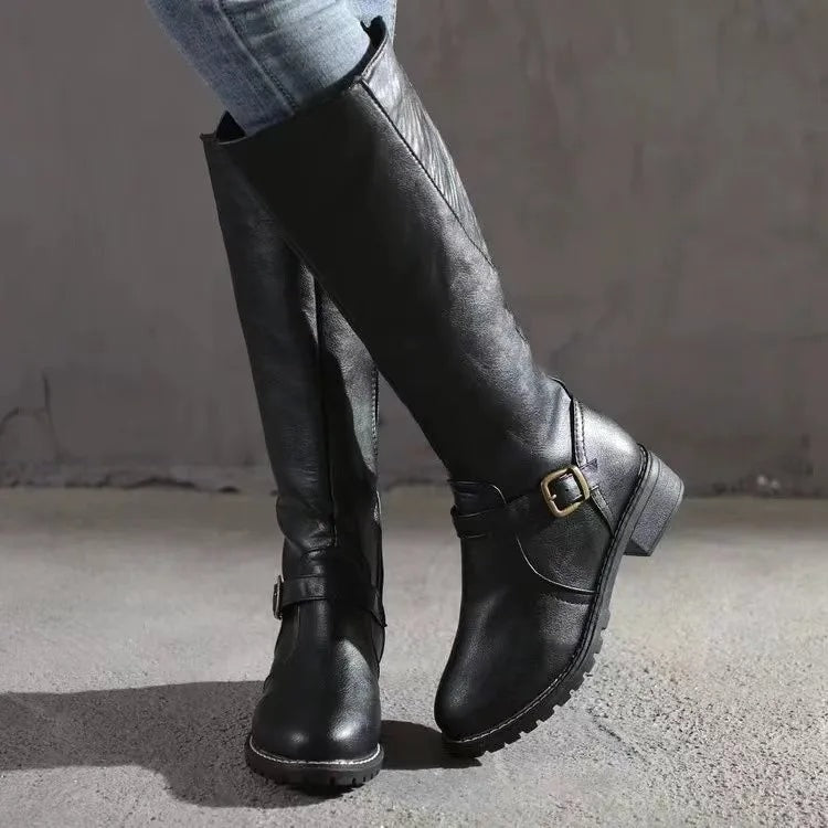 Black knee-high boots shown in a crossed-leg view, with ankle buckles, low block heels, and textured tread soles.
