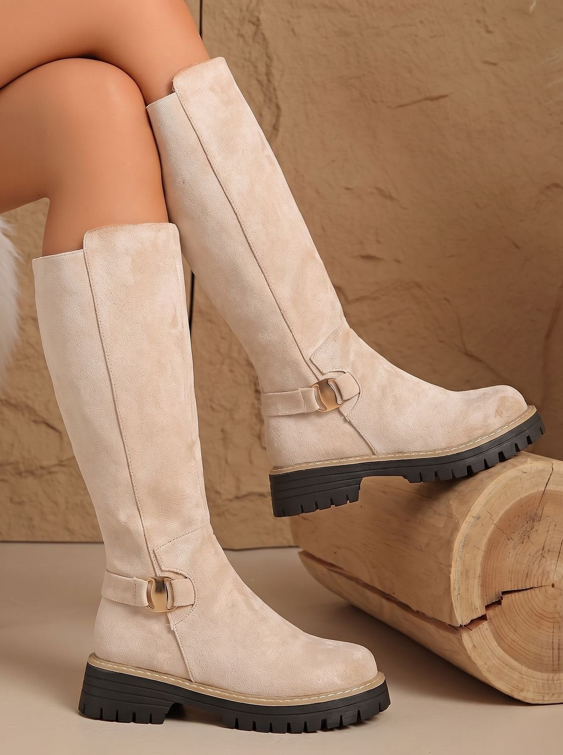 Cream knee-high boots resting against a log, featuring decorative buckles, low block heels, and thick black treaded soles.