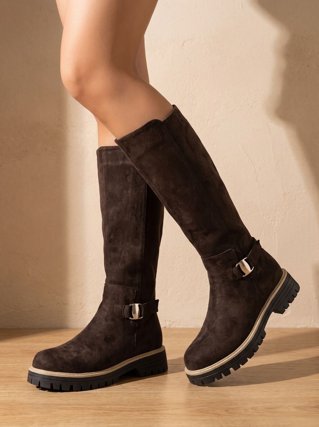 Brown knee-high boots worn against a sunlit wall, with rounded toes, decorative buckles, low heels, and textured lug soles.