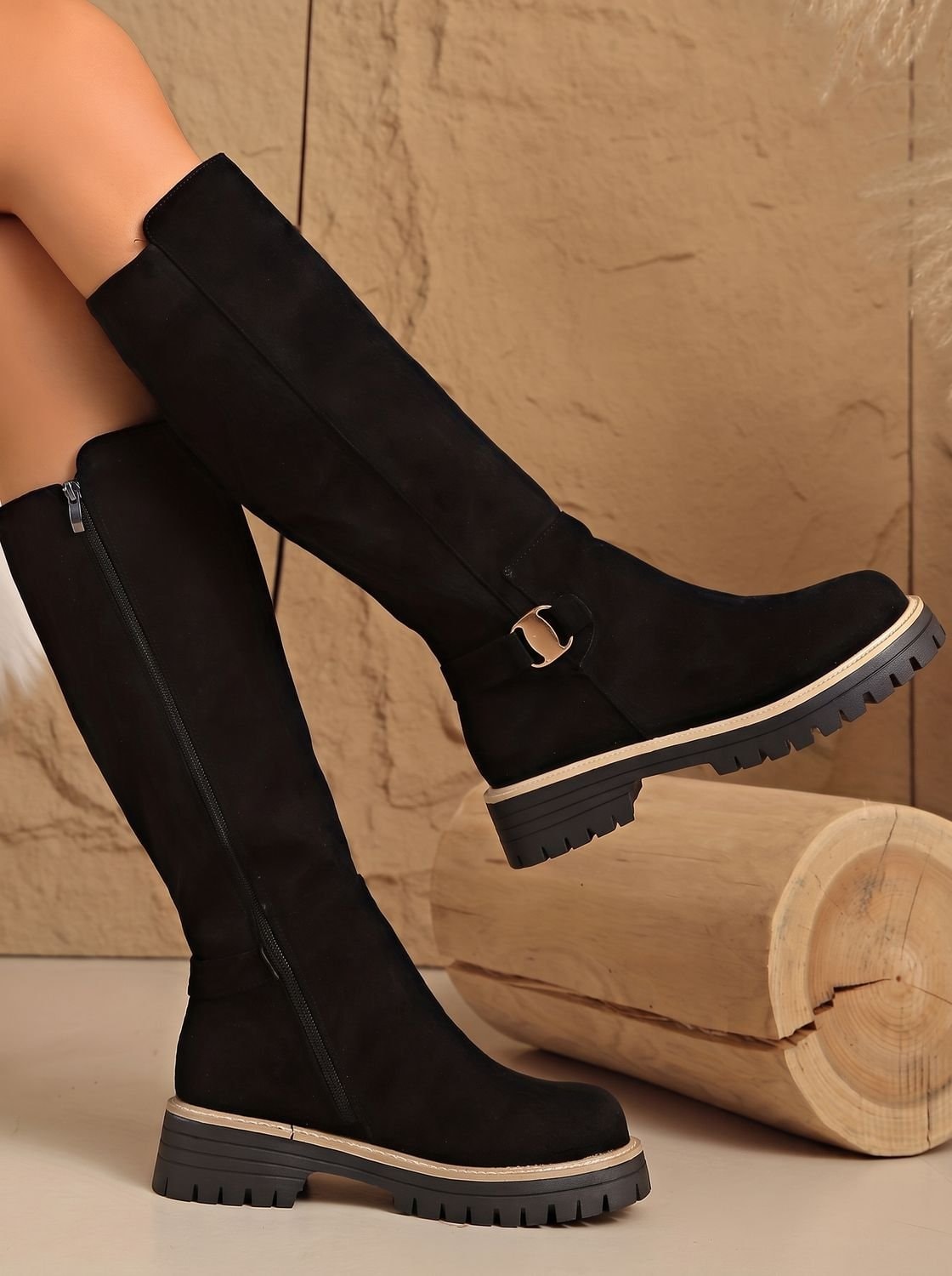 Black knee-high boots posed beside a log, with one boot lifted to reveal the decorative buckle and deep outsole grooves.