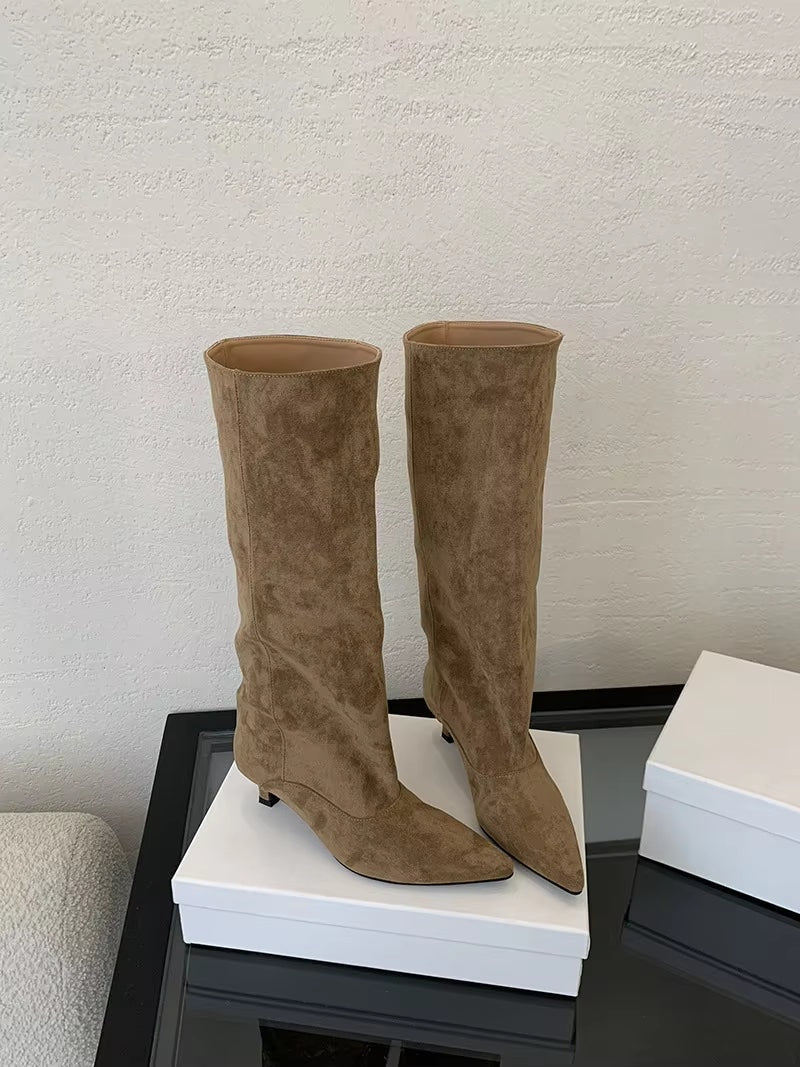 Tan pointed-toe knee-high boots arranged on stacked white boxes, viewed against a textured wall.