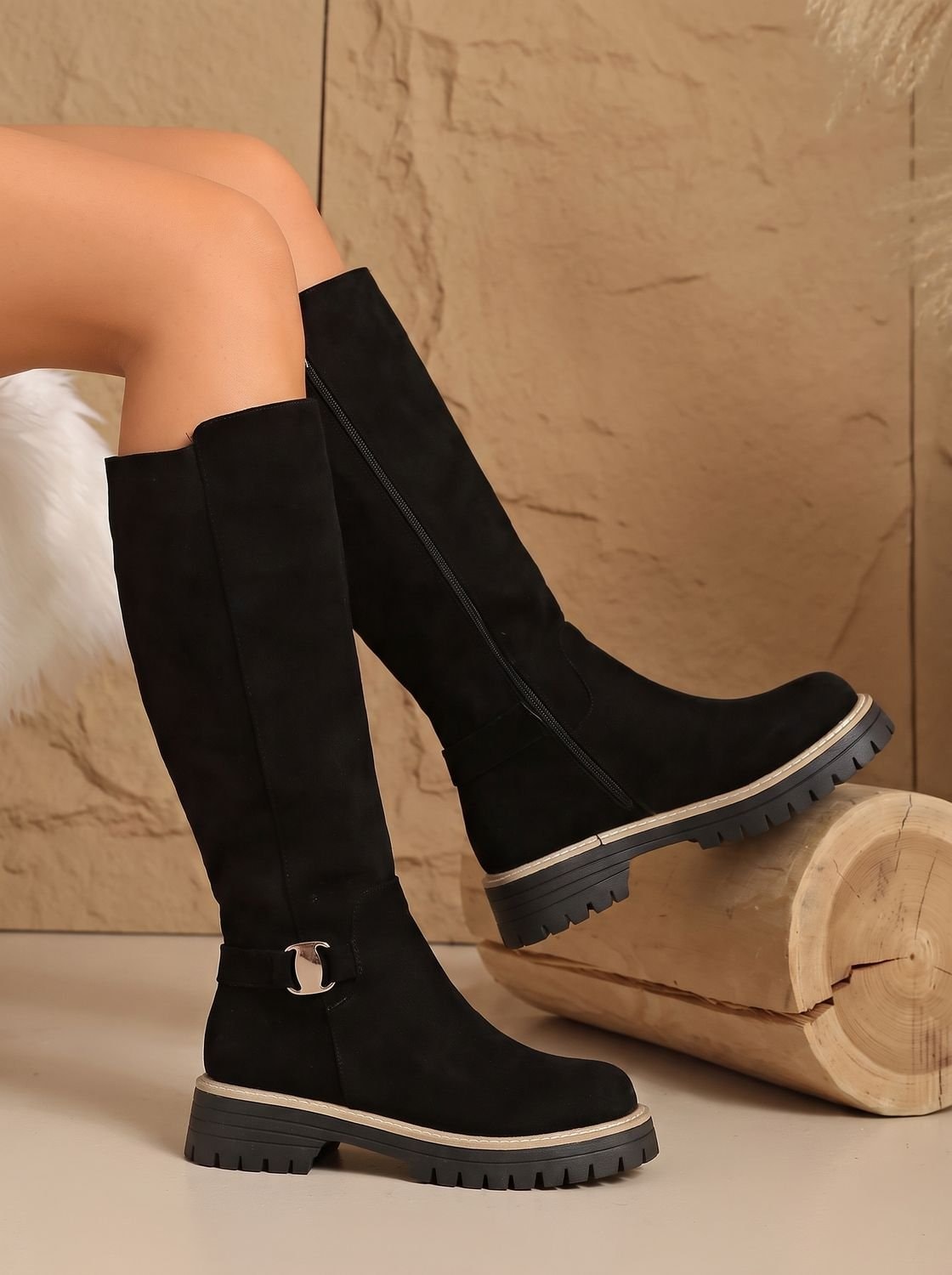Black knee-high boots with side zippers, ankle buckles, contrasting welt trim, and chunky treaded soles, shown beside a log.
