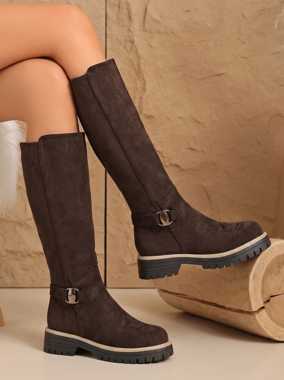 Brown knee-high side-zip boots with metallic ankle buckles and chunky lug soles, shown in a raised-leg pose.