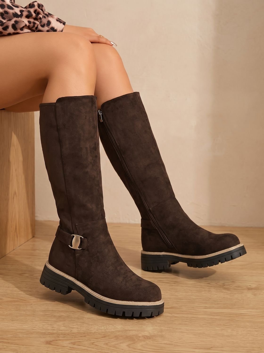 Brown knee-high boots styled on a seated wearer, showing their side zippers, ankle buckles, and chunky soles.
