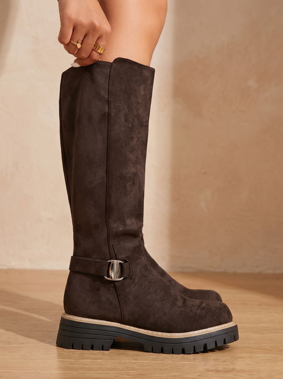 Close side view of a brown knee-high boot with a metallic ankle buckle, tall shaft, contrasting welt trim, and lug sole.