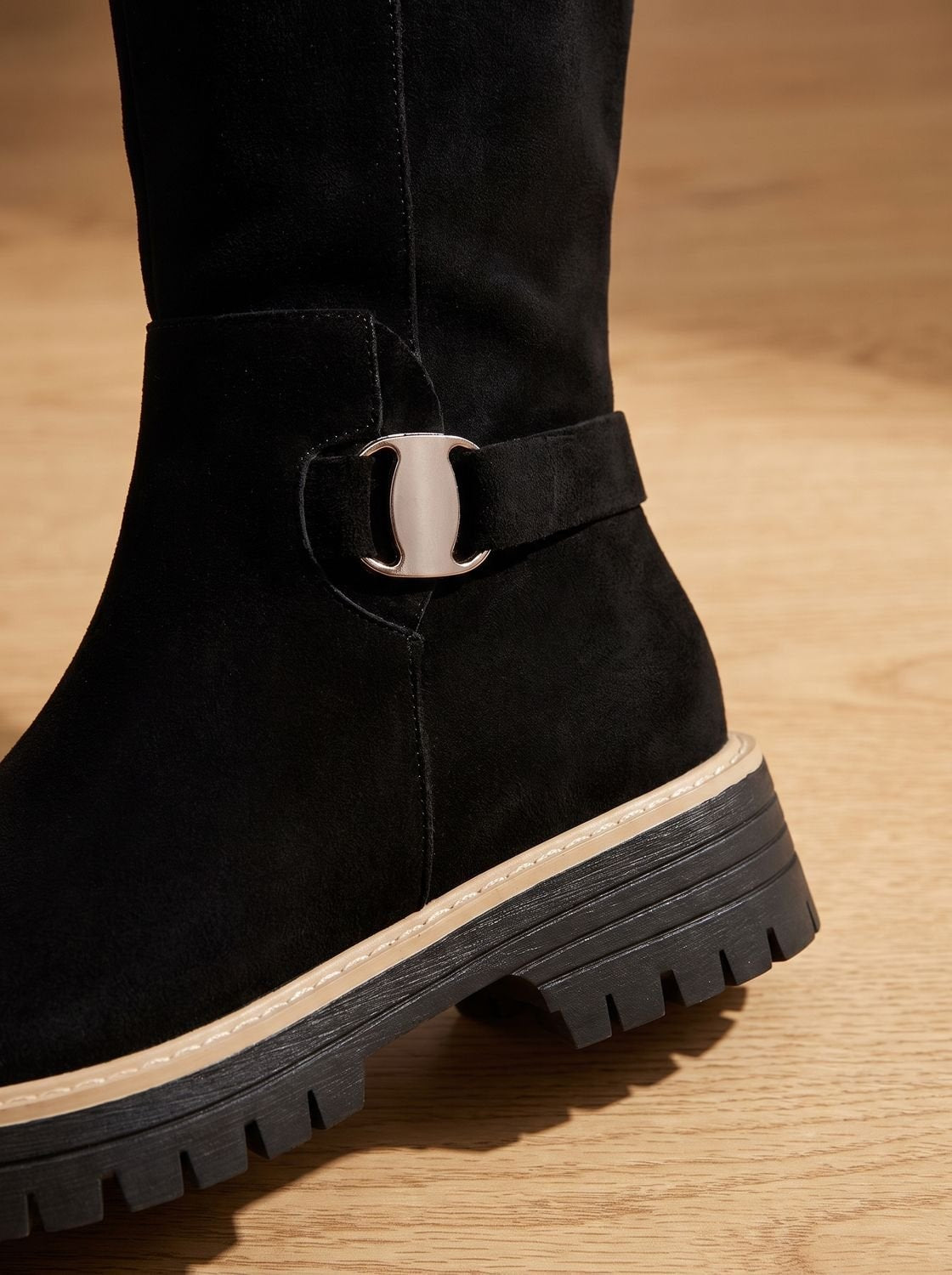 Close-up of a black boot showing its metallic ankle buckle, side seam, contrasting welt trim, and deep lug sole.