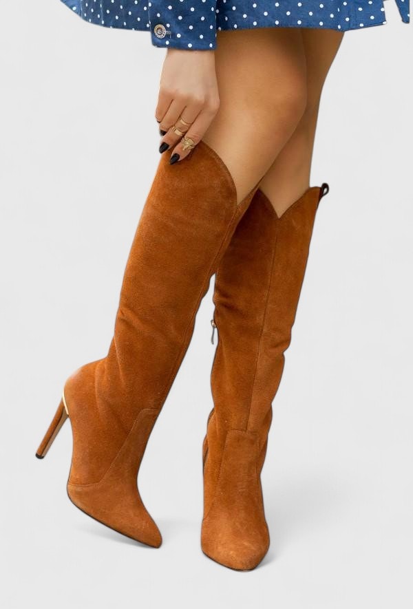 Brown knee-high pointed-toe boots shown with one leg crossed, revealing the tall shafts and side zip.