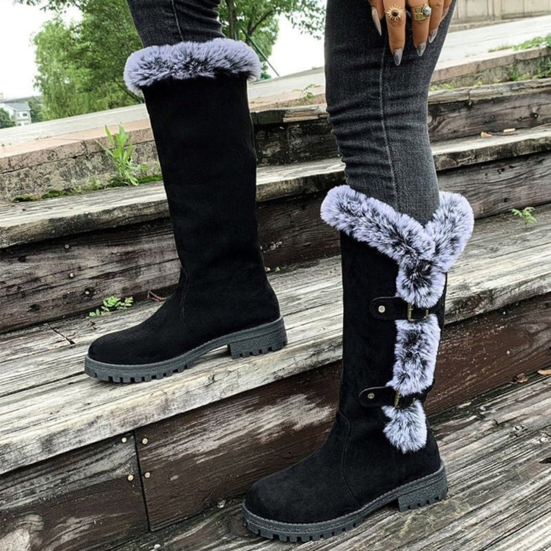 Black knee-high winter boots with contrasting faux fur cuffs, side buckle straps, low block heels, and lug soles shown on wooden steps.