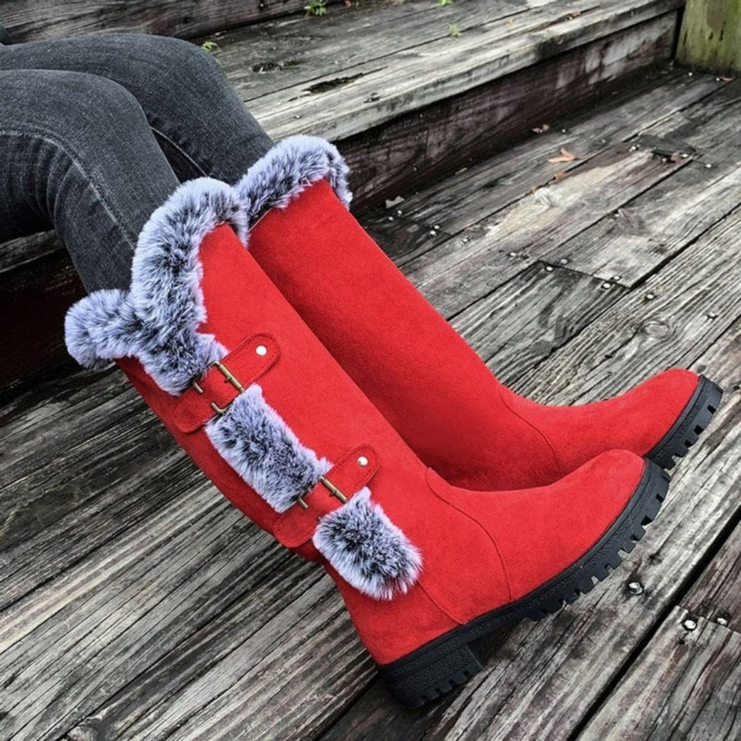 Red knee-high winter boots lying on wooden decking, with faux fur cuffs, side buckle straps, low heels, and rugged lug soles visible.