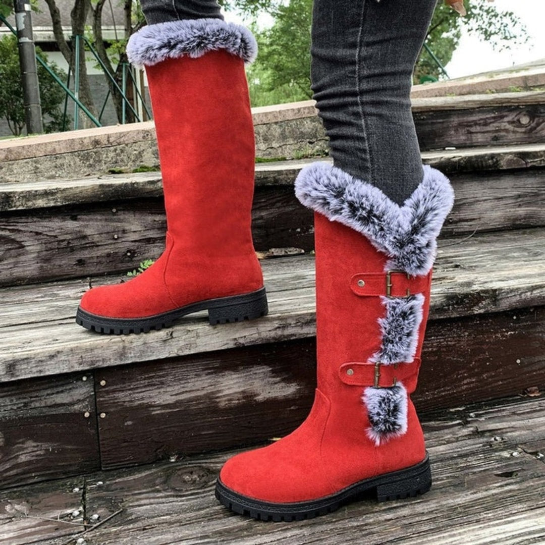 Red winter boots standing on steps in side view, showing the tall silhouette, bright faux fur trim, two buckle straps, and low block heels.