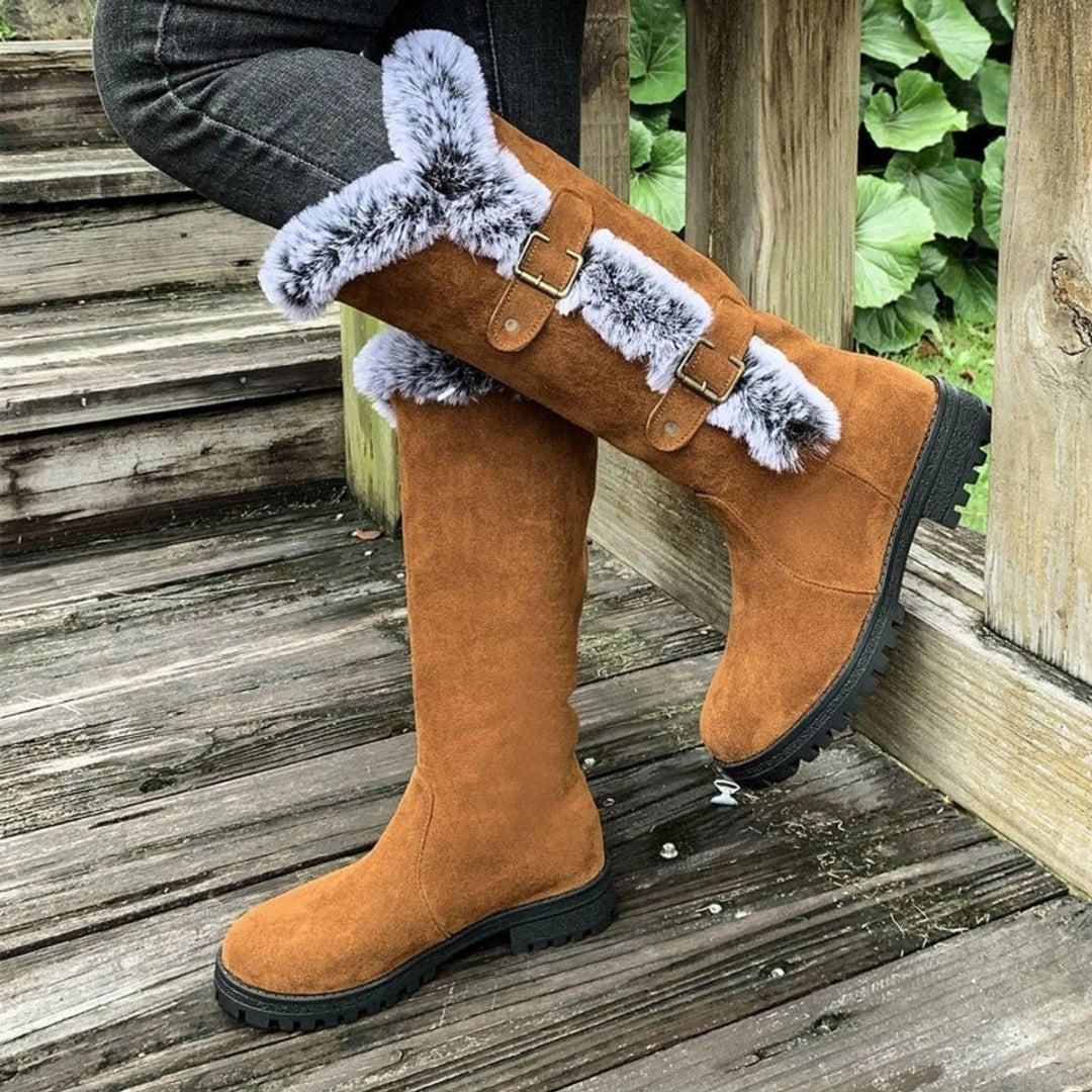 Tan knee-high winter boots shown at an angle, with faux fur trim crossing the outer shaft, twin buckles, and black lug soles.