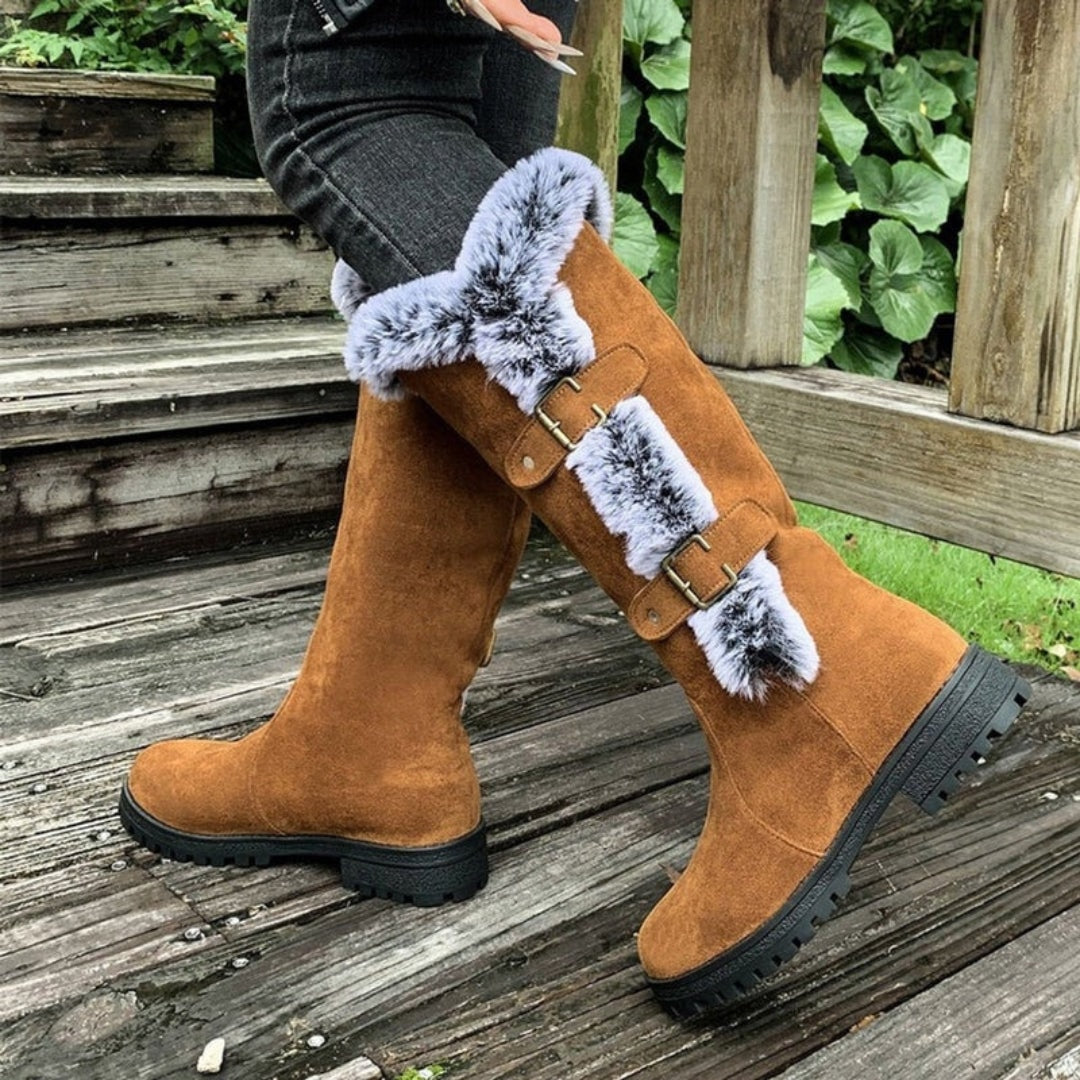 Pair of tan knee-high boots worn with dark jeans, showing their extended shafts, faux fur side accents, buckle closures, and textured soles.