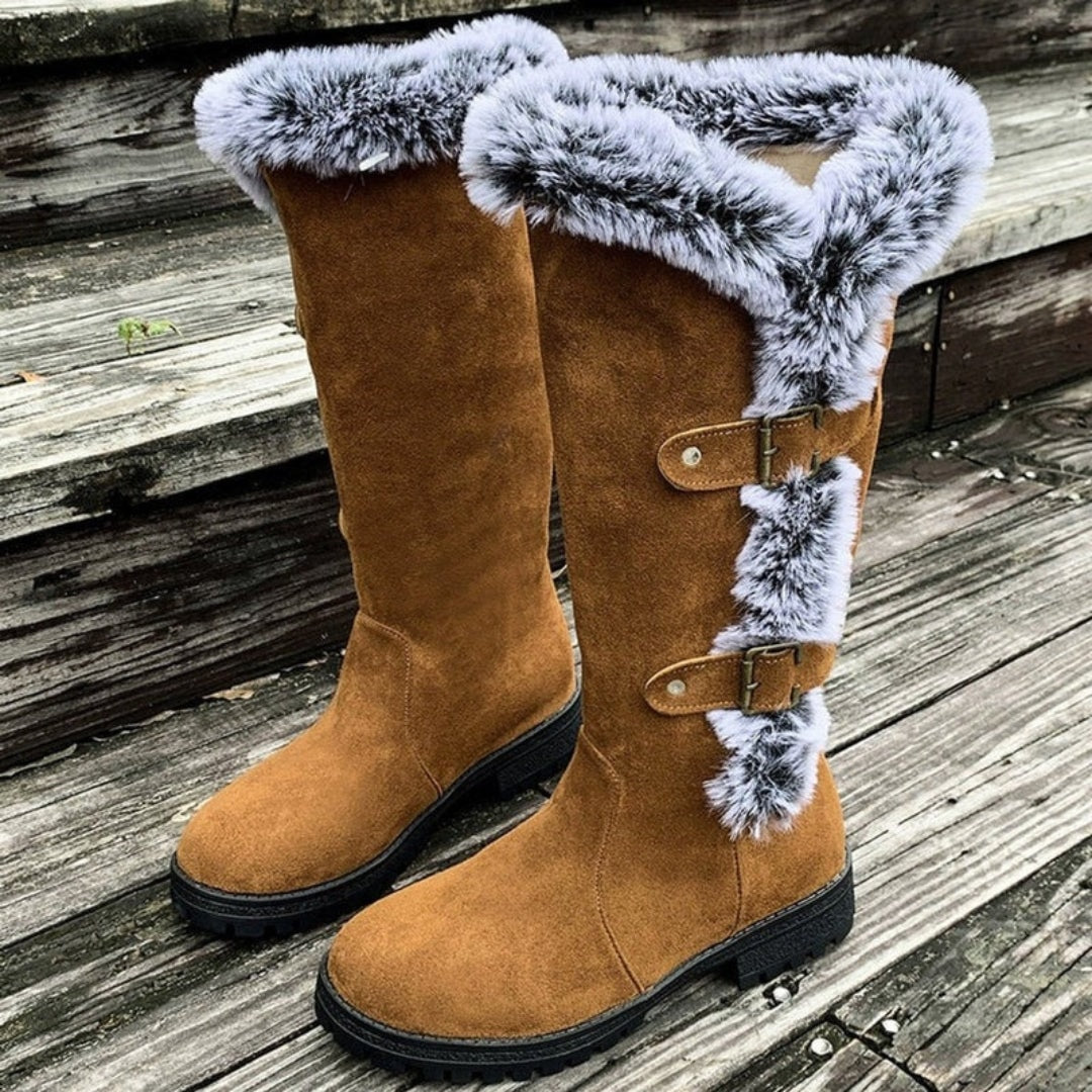 Tan tall winter boots posed upright, with one boot showing the full faux fur cuff and vertical buckle detail along its outer side.