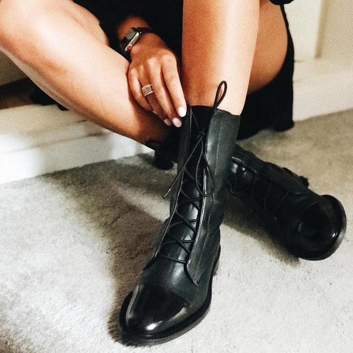 One black lace-up boot being adjusted by hand while the matching boot rests behind it on carpet.