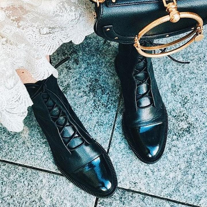 Black lace-up boots styled with a light outfit and handbag, displayed diagonally on textured pavement.