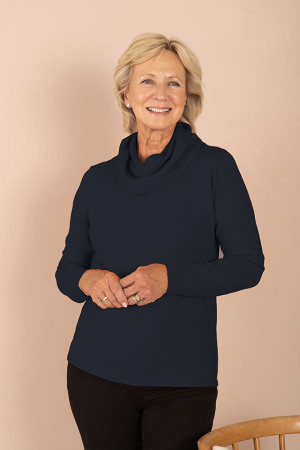 Woman wearing a long sleeve cowl neck top with ribbed cuffs and a centre front seam, with dark trousers._0
