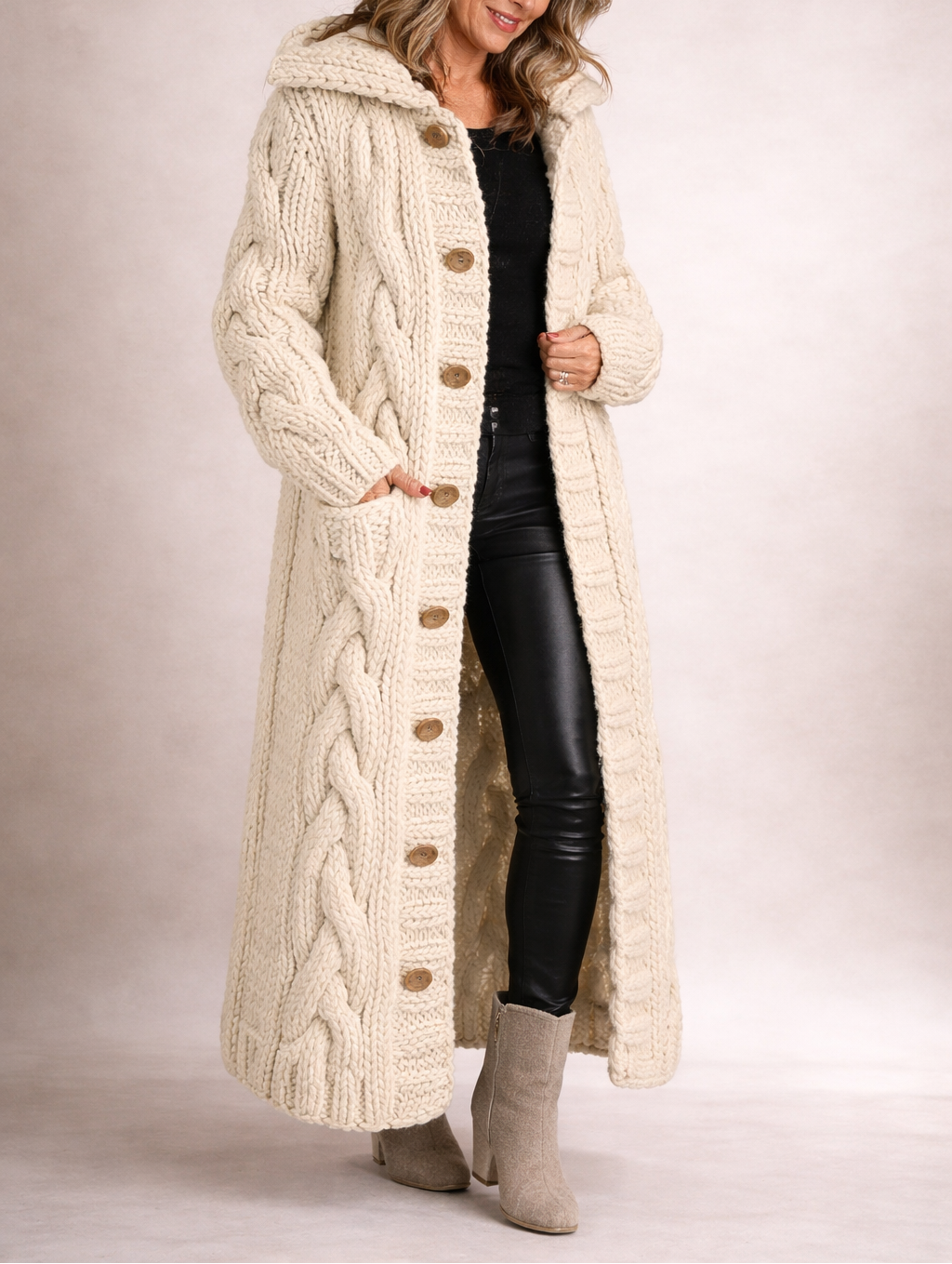 Cream long cable-knit hooded cardigan coat shown with its button front fastened over a black outfit and hands tucked into the side pockets.