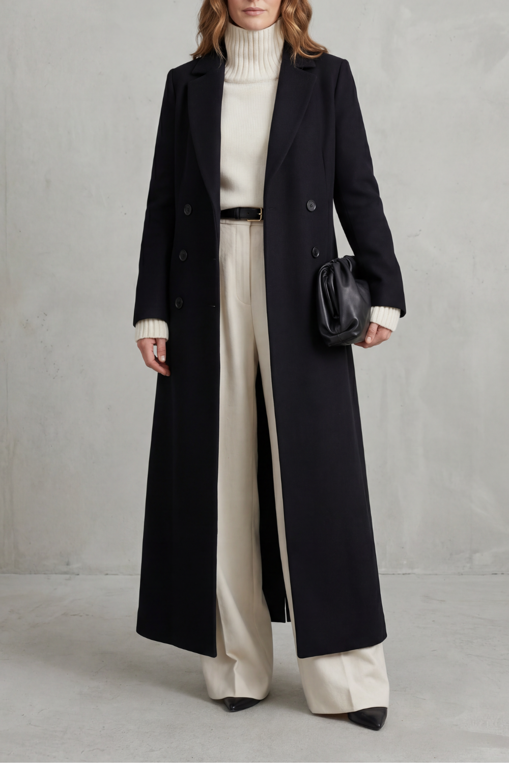 Front view of a black long double-breasted tailored coat with notched lapels and long sleeves, styled over a cream turtleneck and trousers.