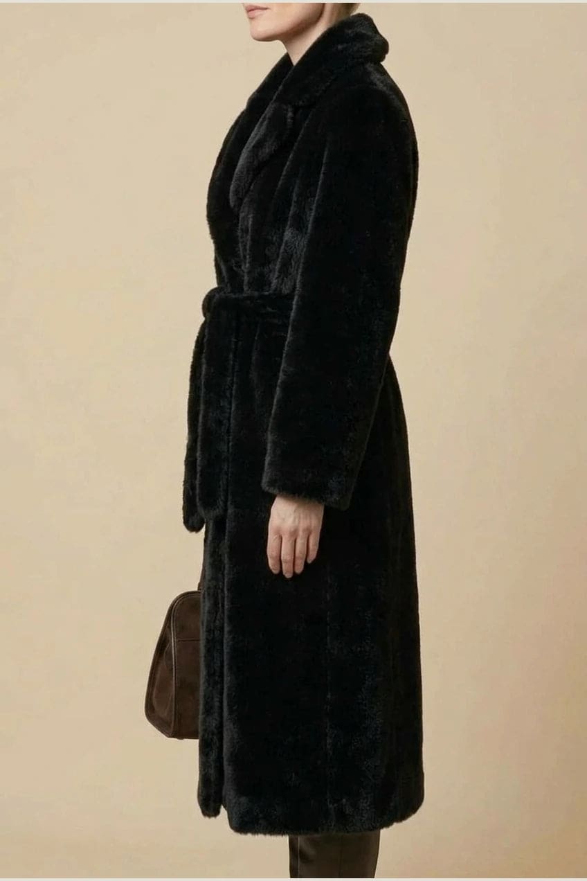 Woman wearing a long belted faux fur coat with notch lapels, tie waist and full-length sleeves, holding a suede tote and wearing knee-high boots._11