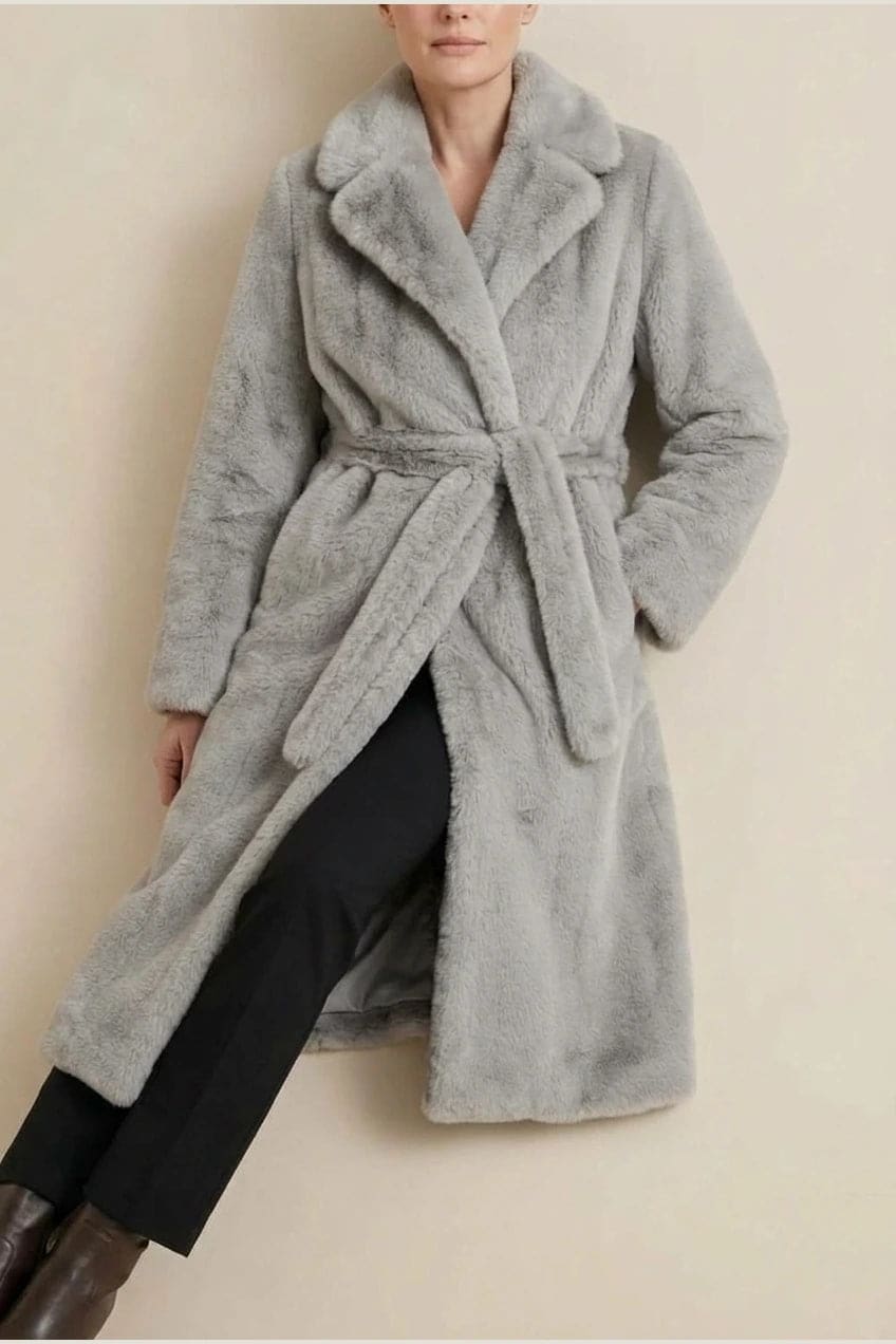 Woman wearing a long belted faux fur coat with notch lapels, tie waist and full-length sleeves, holding a suede tote and wearing knee-high boots._15