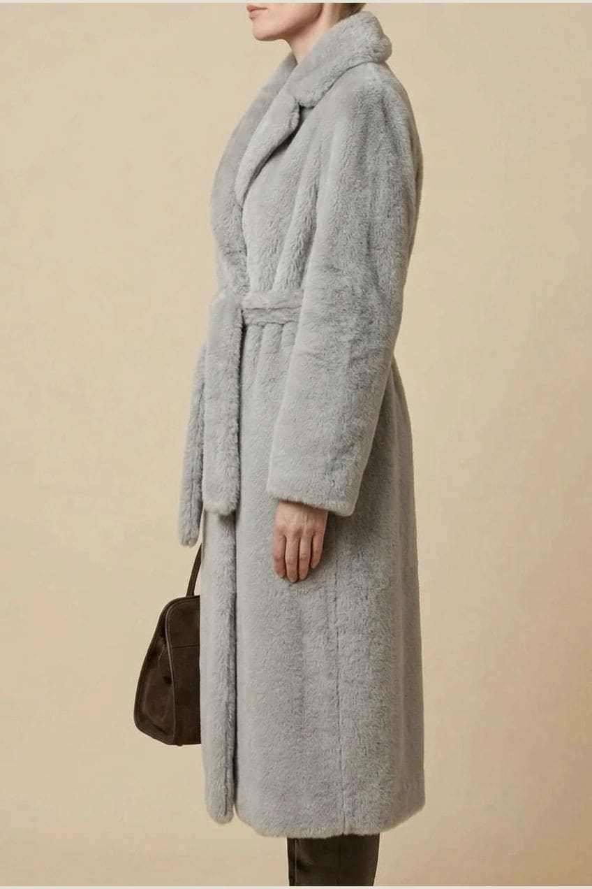 Woman wearing a long belted faux fur coat with notch lapels, tie waist and full-length sleeves, holding a suede tote and wearing knee-high boots._17