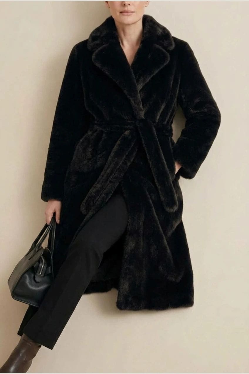 Woman wearing a long belted faux fur coat with notch lapels, tie waist and full-length sleeves, holding a suede tote and wearing knee-high boots._9