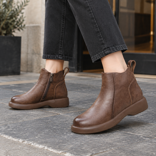 Brown ankle boots with rounded toes, side zippers, rear pull tabs, and low wedge soles shown with black cropped trousers.