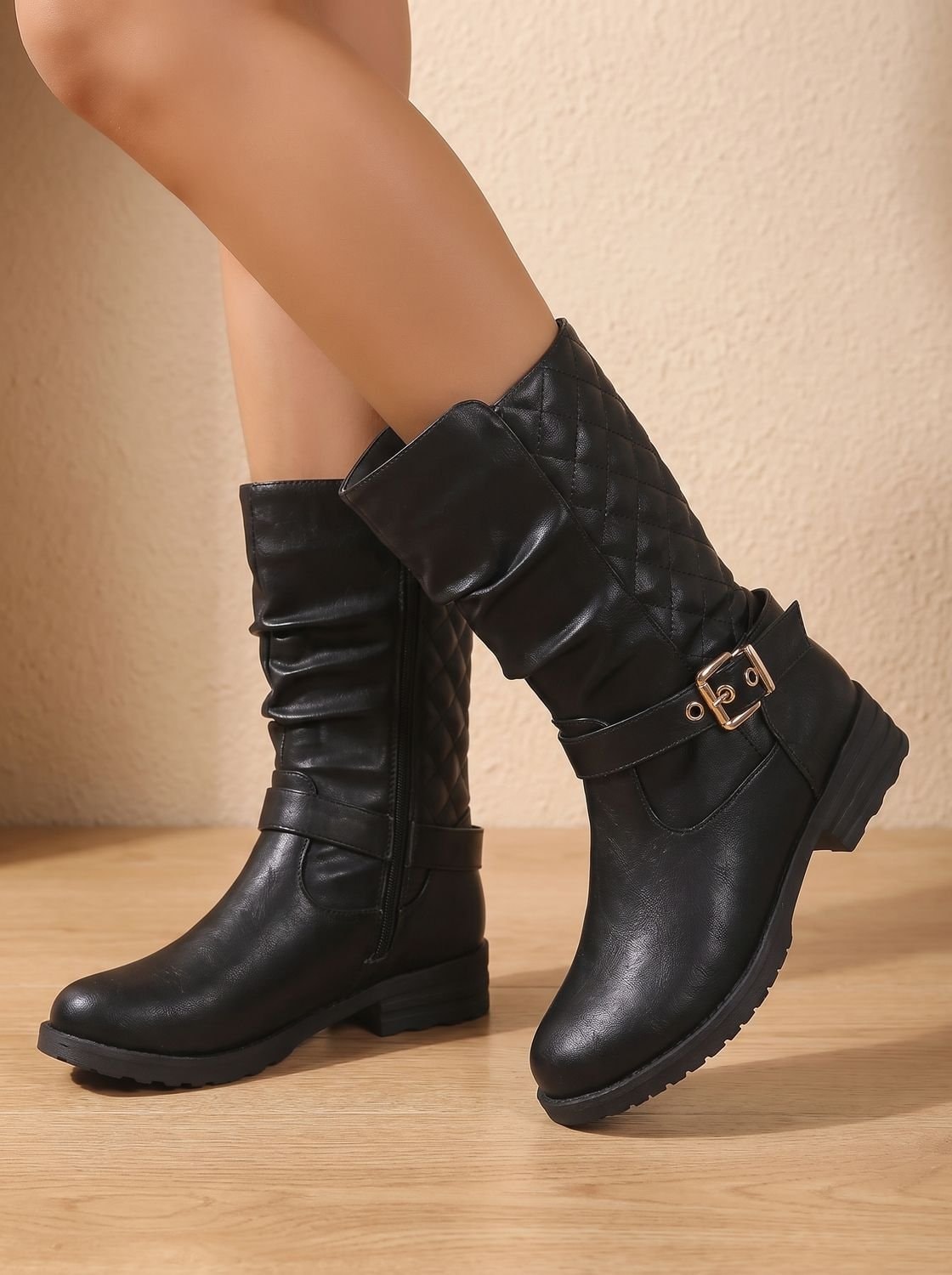 Angled view of black mid-calf boots with ruched fronts, quilted outer shafts, buckle accents, and rounded toes.