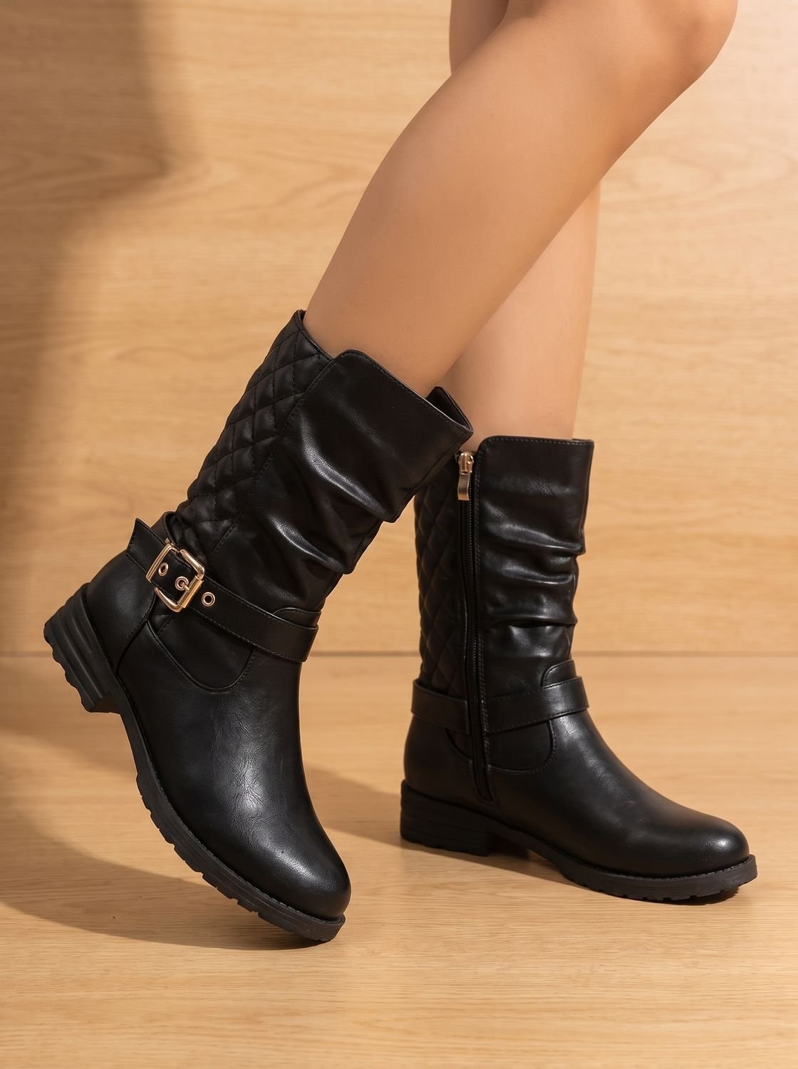 Pair of black boots on a wood floor, with one raised to reveal the side zipper, buckle strap, and low heel.