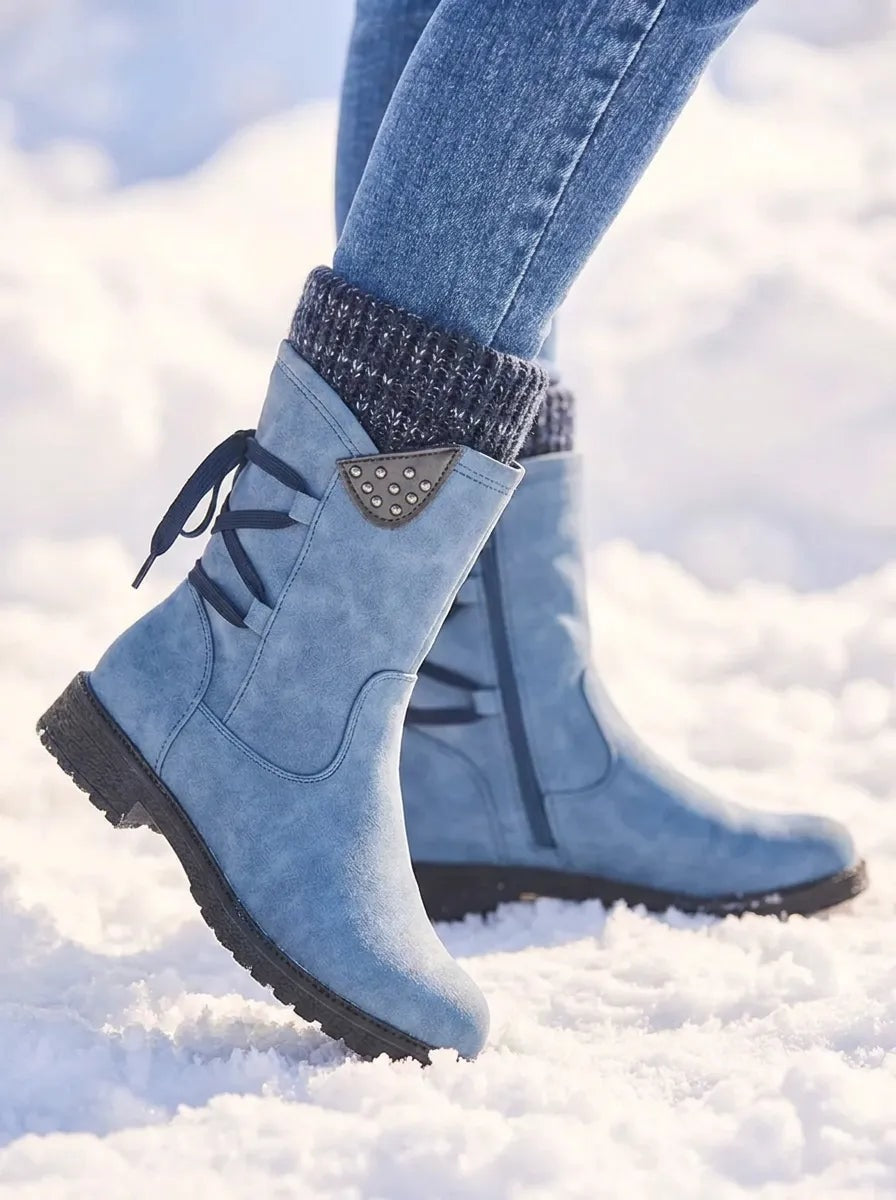 Side view of a light blue winter boot lifted above the snow, showing its treaded sole, knitted cuff, side zip and decorative studded detail.