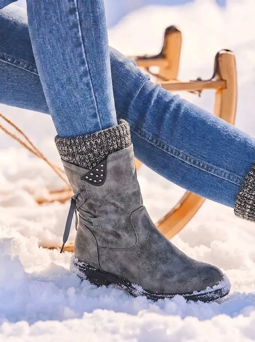 Grey mid-calf winter boot in snow with a contrasting knitted cuff, decorative studded side tab and dark treaded sole.