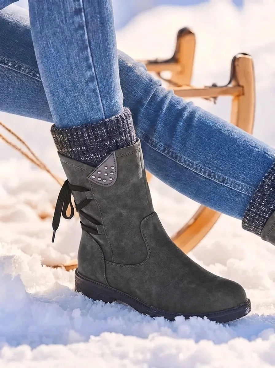 Black mid-calf winter boot shown in profile against snowy ground, with a dark knitted cuff, side zip and rugged sole.