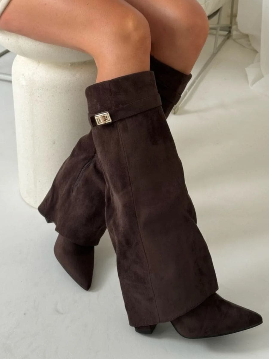 Dark brown pointed-toe knee-high boots styled while seated, with the extended fold-over silhouette and metallic cuff detail visible.