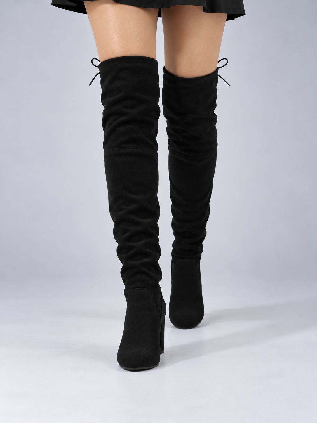 Front view of black over-the-knee boots worn beneath a short skirt, emphasizing their tall ruched shafts and rounded toes.