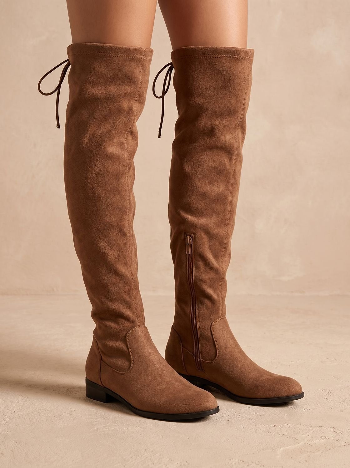 Brown over-the-knee flat boots shown from the side with visible back ties, rounded toes, and a full-length side zip.