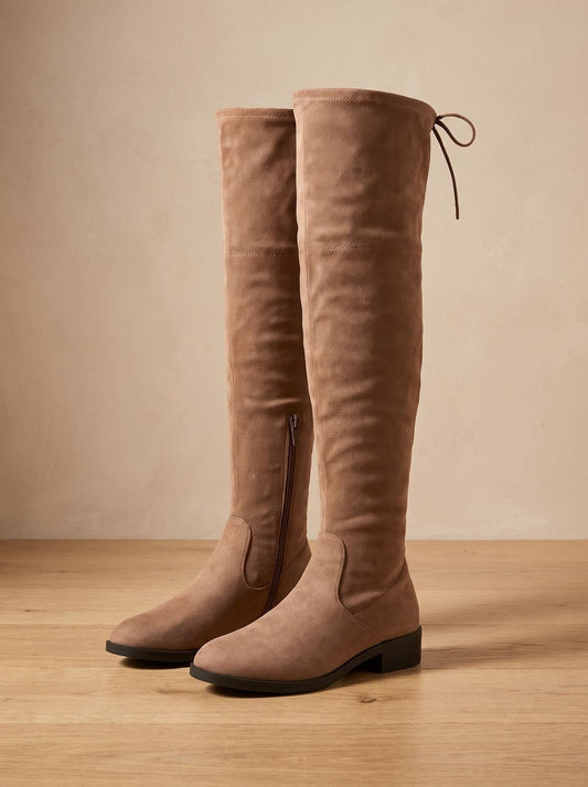 Tan over-the-knee flat boots displayed in a front three-quarter view, highlighting the low block heels, rounded toes, and tall fitted shafts.