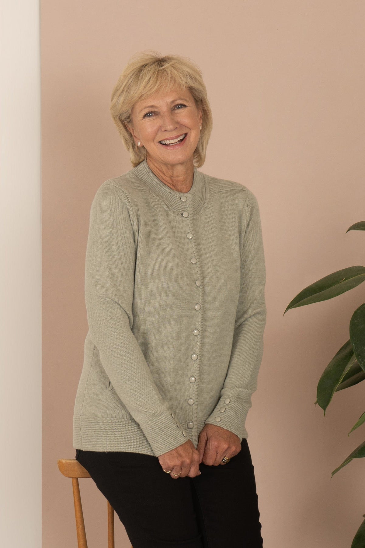 Woman wearing a long sleeve knitted cardigan with a ribbed funnel neck, full button front, side pockets and buttoned cuffs, with dark trousers._0
