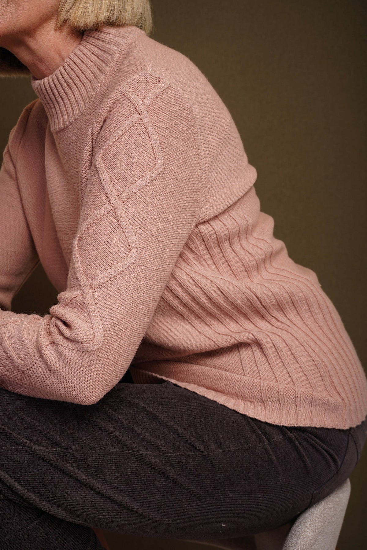 Woman sitting and smiling, wearing a relaxed cable knit turtleneck jumper with ribbed cuffs and diamond patterned sleeves, paired with corduroy trousers._15
