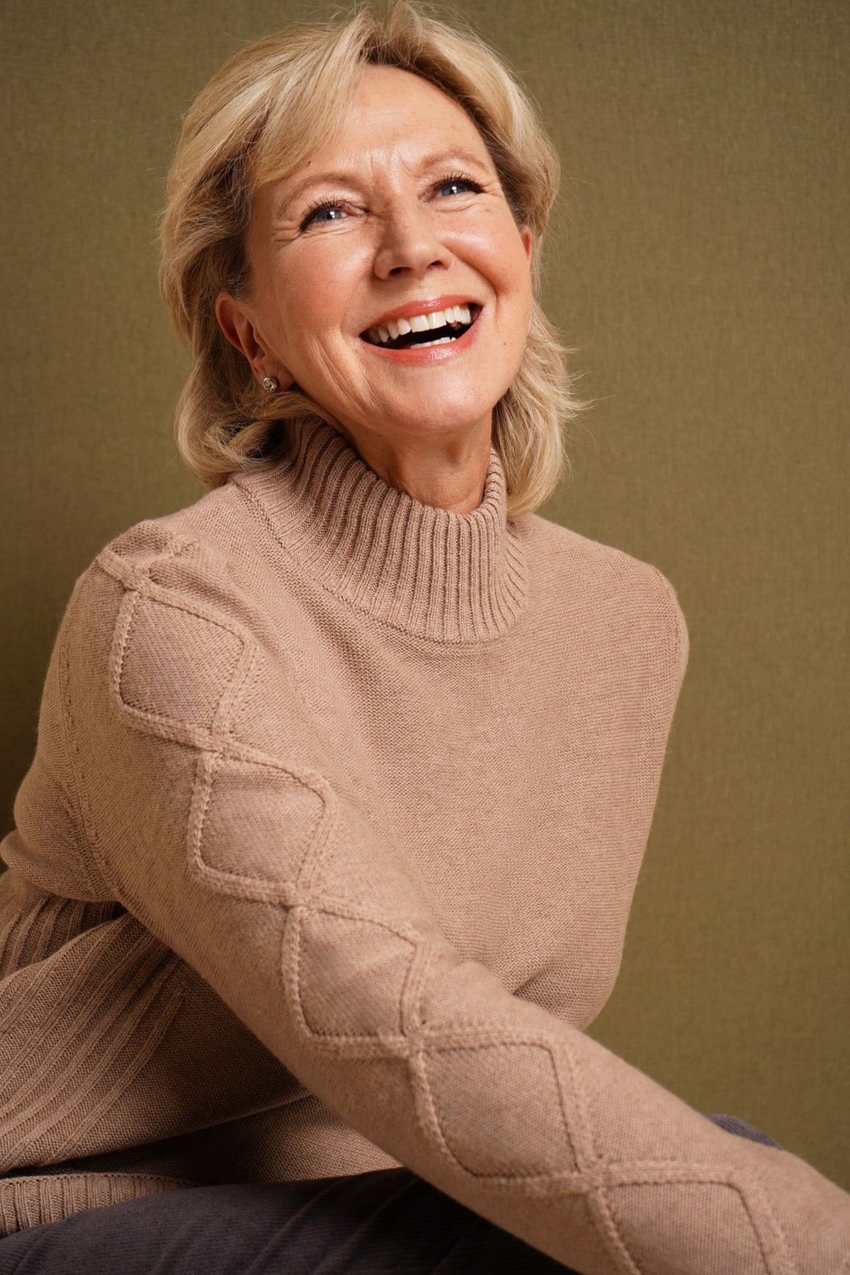 Woman sitting and smiling, wearing a relaxed cable knit turtleneck jumper with ribbed cuffs and diamond patterned sleeves, paired with corduroy trousers._4
