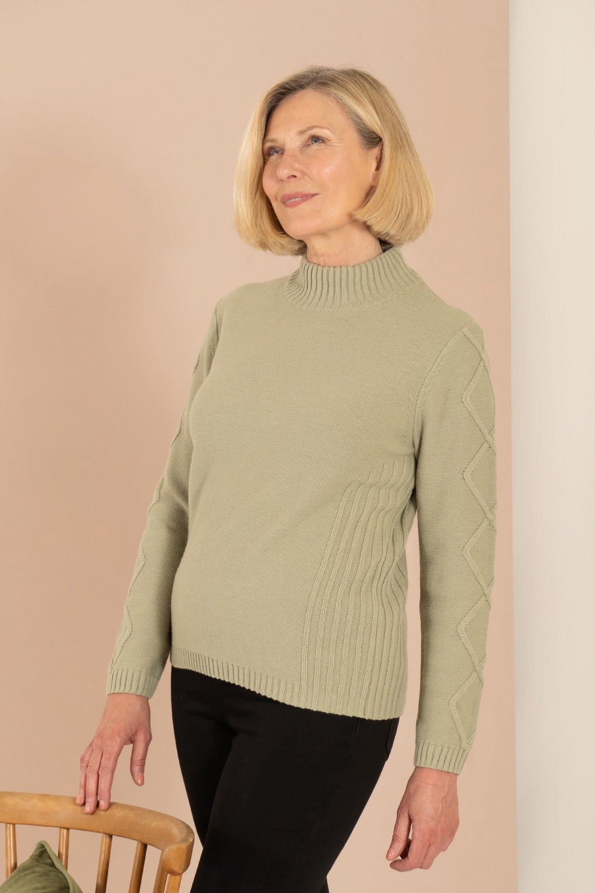 Woman sitting and smiling, wearing a relaxed cable knit turtleneck jumper with ribbed cuffs and diamond patterned sleeves, paired with corduroy trousers._7