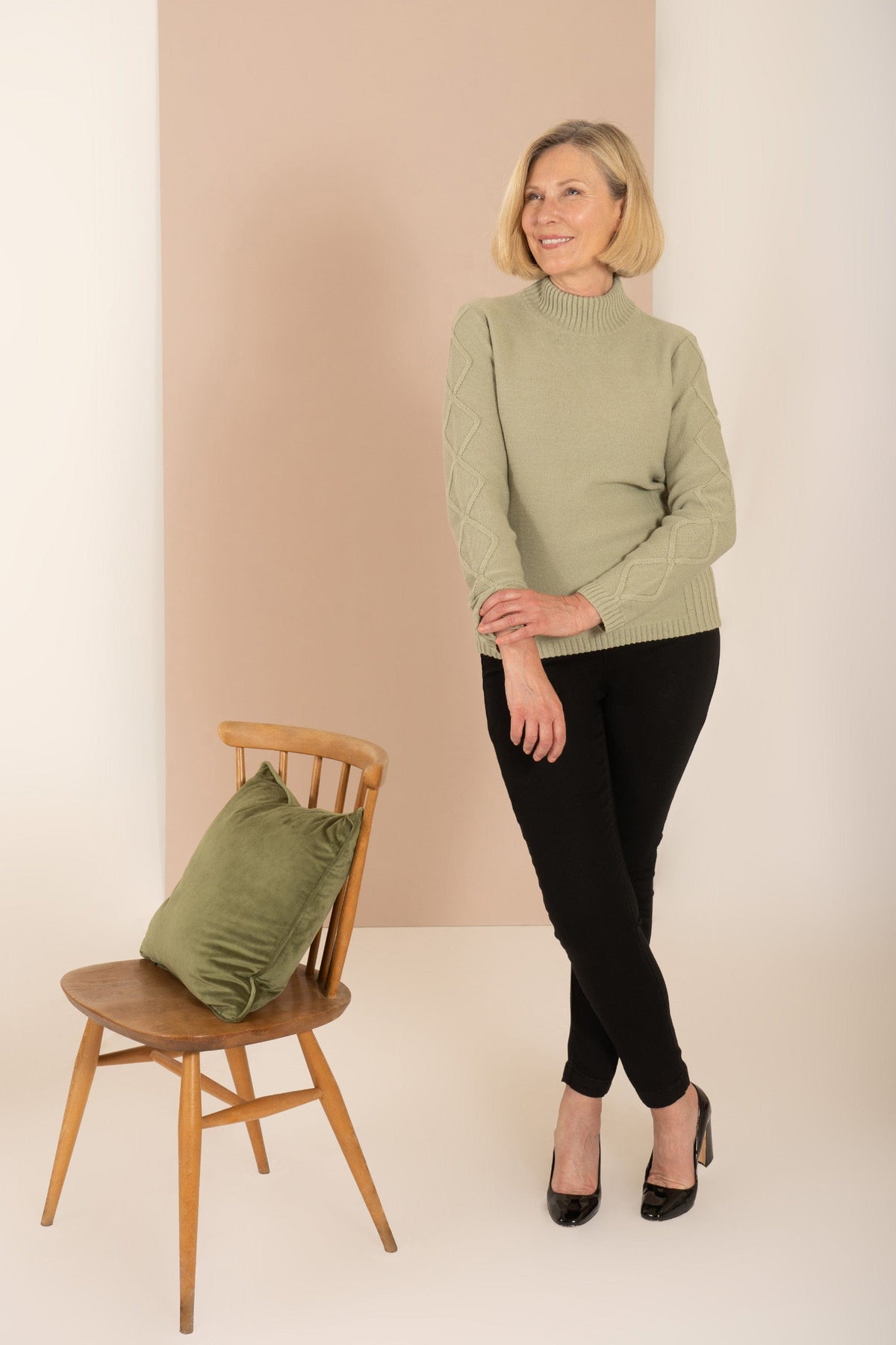 Woman sitting and smiling, wearing a relaxed cable knit turtleneck jumper with ribbed cuffs and diamond patterned sleeves, paired with corduroy trousers._8