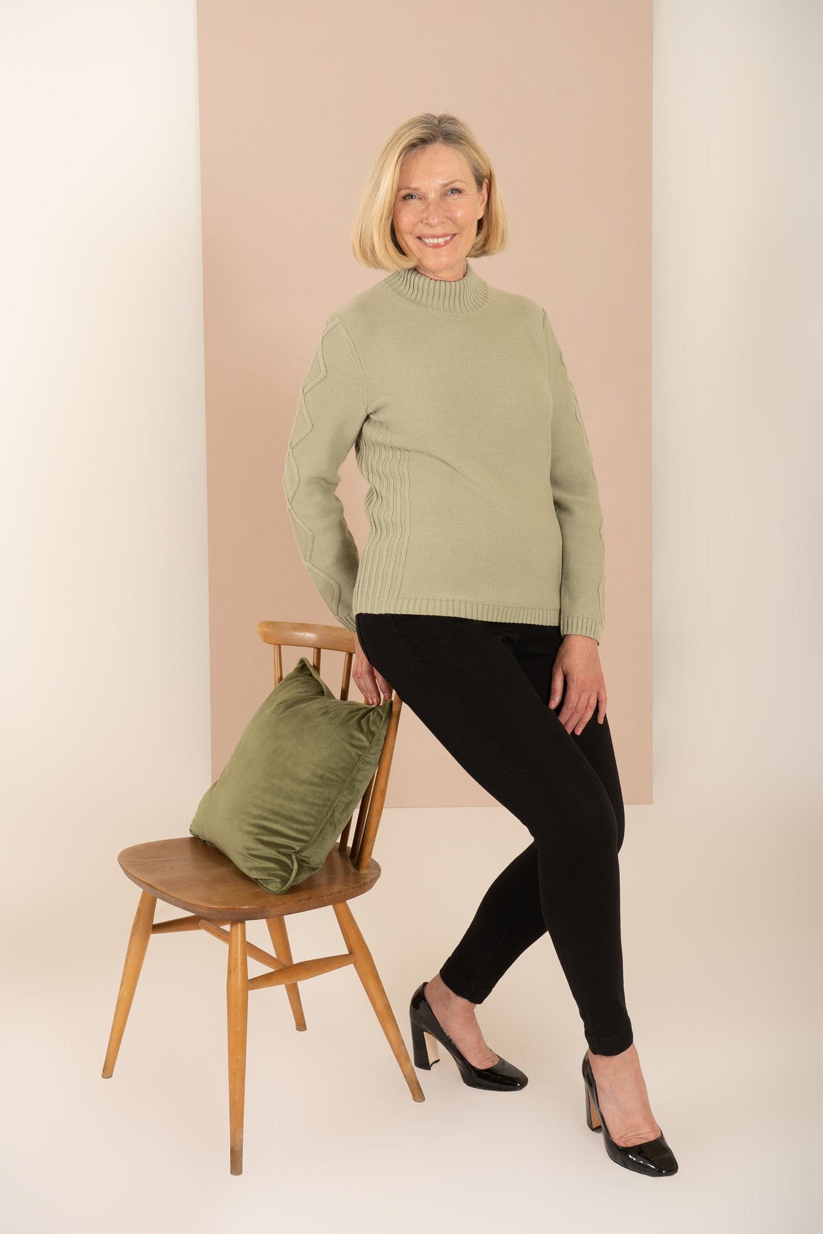 Woman sitting and smiling, wearing a relaxed cable knit turtleneck jumper with ribbed cuffs and diamond patterned sleeves, paired with corduroy trousers._9