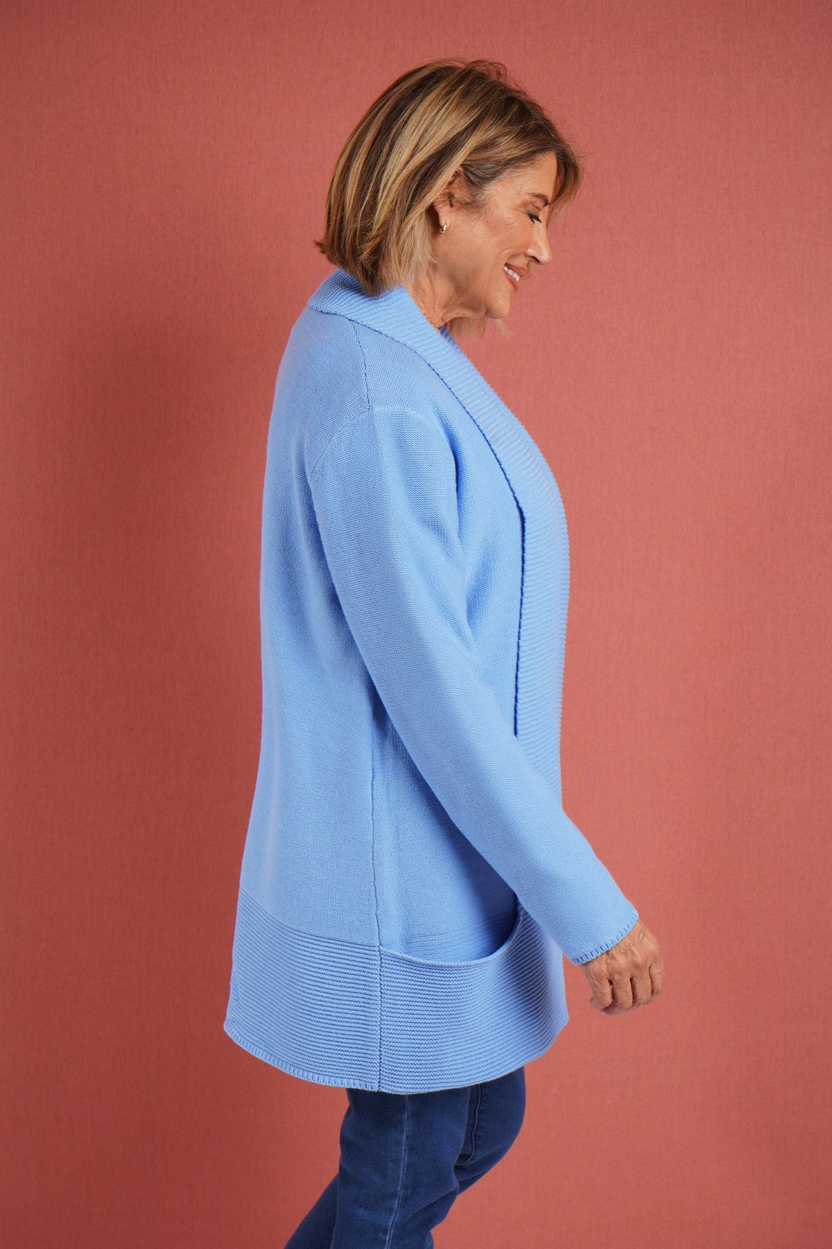 Woman wearing a longline open front knitted cardigan with a wide ribbed shawl collar and front patch pockets over a plain top and jeans._3
