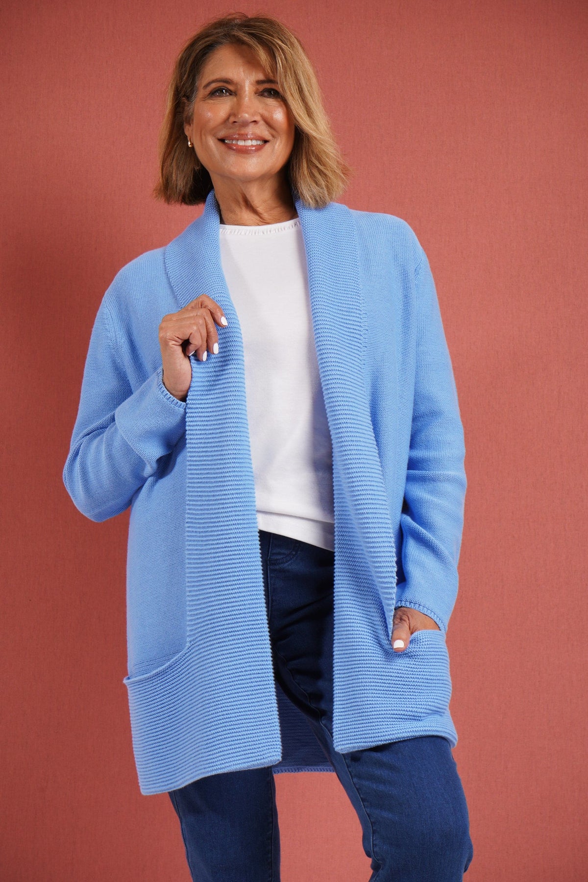 Woman wearing a longline open front knitted cardigan with a wide ribbed shawl collar and front patch pockets over a plain top and jeans._4