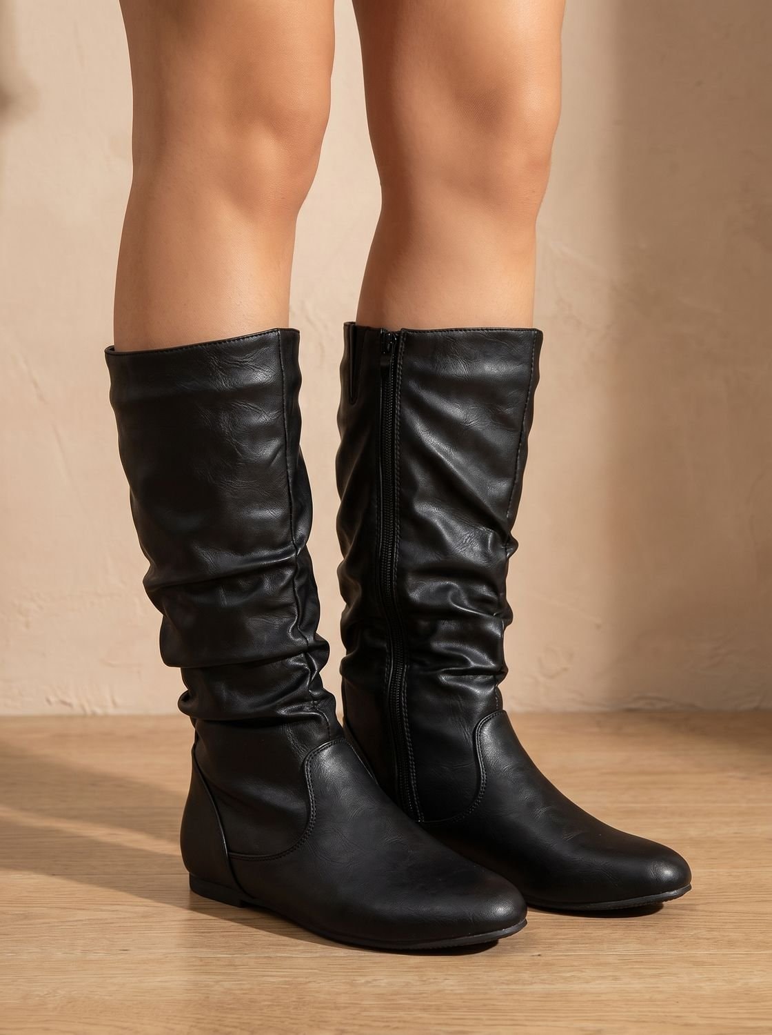 Pair of black knee-high boots worn on bare legs, showing their relaxed gathered shafts, rounded toes, and low flat heels.