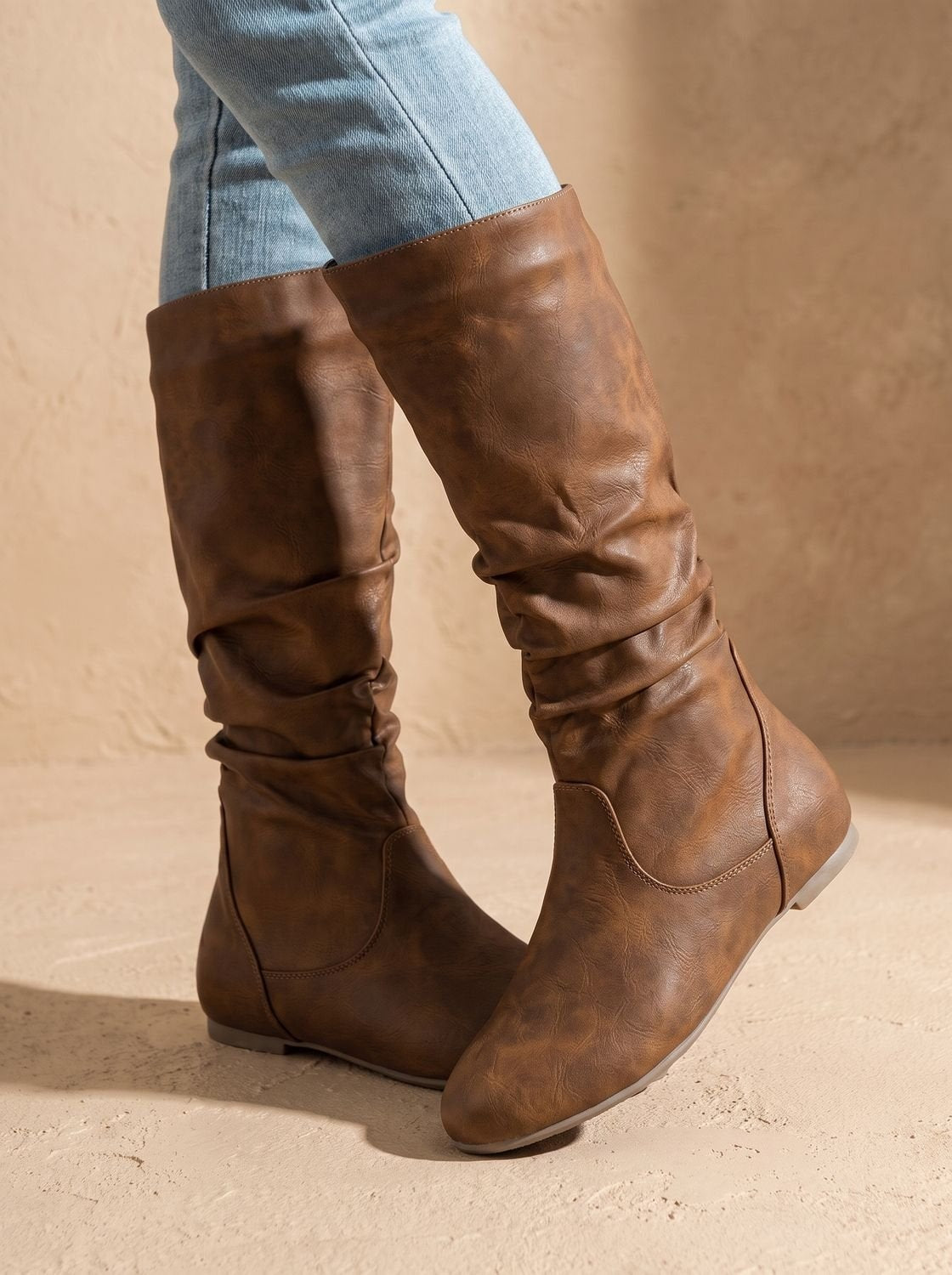 Brown slouchy knee-high boots worn with jeans, shown from the outer side with ruched shafts and rounded toes.